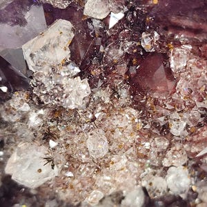 May include: Close-up of a cluster of amethyst crystals. The image showcases a variety of crystal formations, with a deep purple hue dominating the center. Clear, sparkling quartz crystals are interspersed throughout, creating a contrast in color and texture.