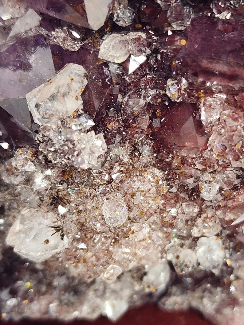 May include: Close-up of a cluster of amethyst crystals. The image showcases a variety of crystal formations, with a deep purple hue dominating the center. Clear, sparkling quartz crystals are interspersed throughout, creating a contrast in color and texture.