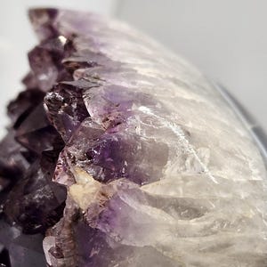 May include: Close-up of an amethyst geode with a crystalline structure. The geode features a gradient of colors, from deep purple to translucent white. The surface is rough and textured, showcasing natural formations.