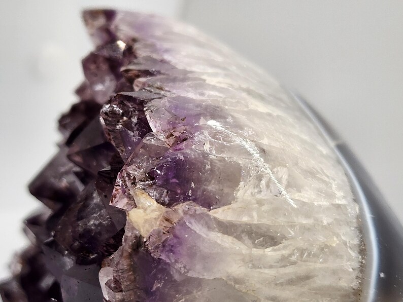 May include: Close-up of an amethyst geode with a crystalline structure. The geode features a gradient of colors, from deep purple to translucent white. The surface is rough and textured, showcasing natural formations.