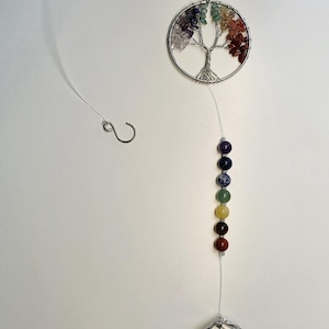 Puede incluir: Un atrapasoles de cristal colgante con un colgante plateado de árbol de la vida, detalles de piedras preciosas de colores y una bola de cristal facetada. El diseño incluye una serie de cuentas de chakra en varios colores, suspendidas de un hilo transparente.