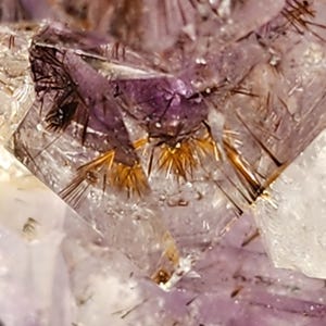 May include: Close-up of a crystal formation with a mix of purple, clear, and gold colors. The crystal has a complex structure with needle-like inclusions and a sparkling appearance. The overall theme is natural gemstones.