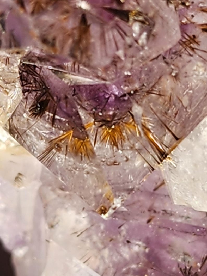 May include: Close-up of a crystal formation with a mix of purple, clear, and gold colors. The crystal has a complex structure with needle-like inclusions and a sparkling appearance. The overall theme is natural gemstones.