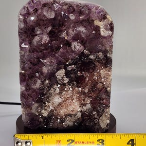 May include: A large amethyst crystal cluster, predominantly purple with white and brown inclusions, mounted on a dark wooden base. The crystal is approximately 4 inches tall, with a rough, textured surface. A yellow measuring tape is placed in front of the crystal.