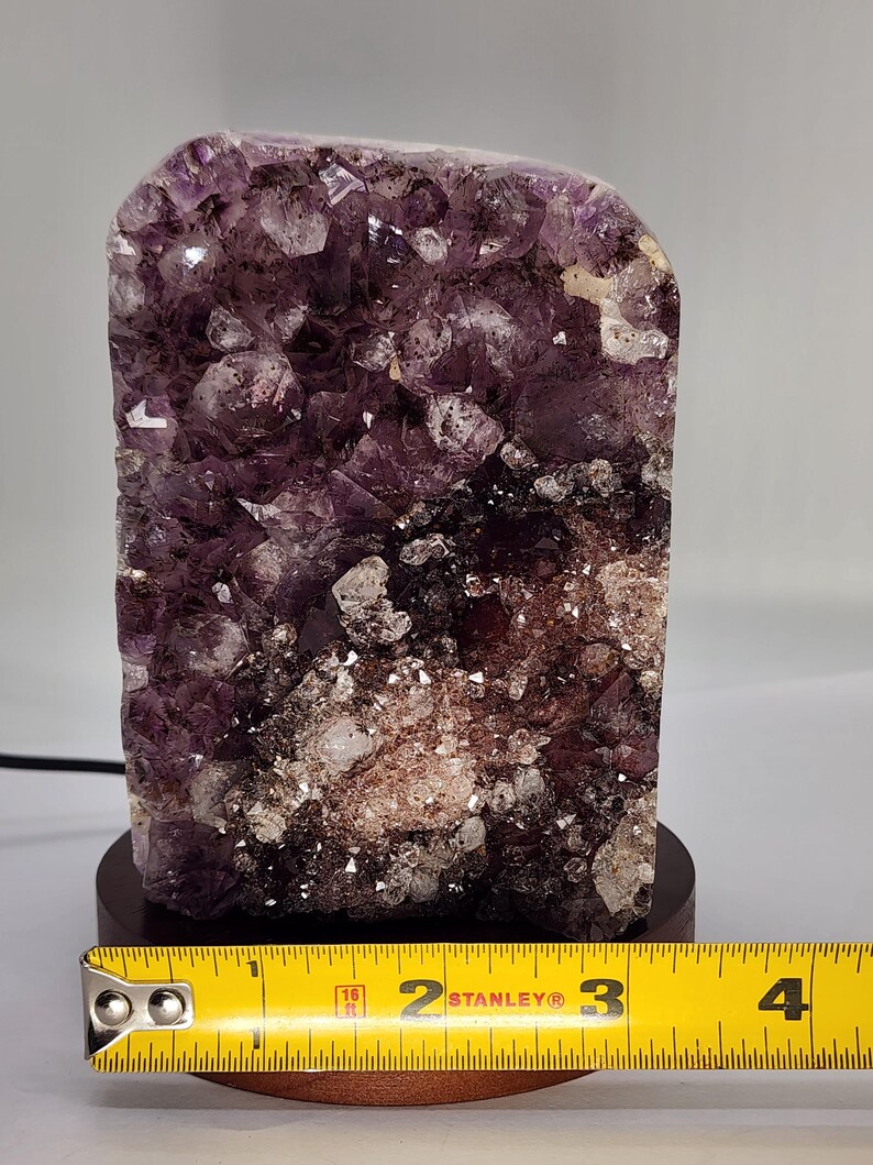 May include: A large amethyst crystal cluster, predominantly purple with white and brown inclusions, mounted on a dark wooden base. The crystal is approximately 4 inches tall, with a rough, textured surface. A yellow measuring tape is placed in front of the crystal.