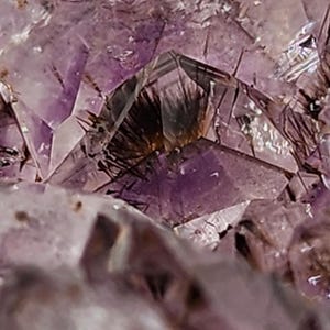 May include: Close-up of a purple amethyst crystal cluster. The crystalline structure features sharp, angular facets and a range of purple hues, with darker inclusions. The natural formation displays a complex, textured surface, showcasing the mineral's geological beauty.