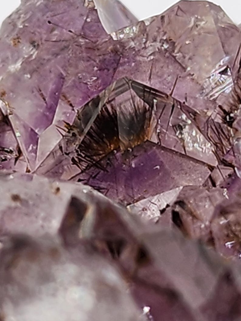 May include: Close-up of a purple amethyst crystal cluster. The crystalline structure features sharp, angular facets and a range of purple hues, with darker inclusions. The natural formation displays a complex, textured surface, showcasing the mineral's geological beauty.