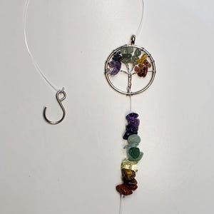 May include: A hanging suncatcher with a silver-colored tree of life pendant, colorful gemstone beads, and a faceted crystal ball. The gemstones are arranged in a rainbow of colors. The suncatcher hangs from a clear string with a metal hook.