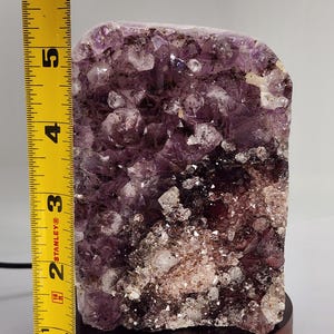 May include: A large amethyst crystal cluster, approximately 5 inches tall, mounted on a round wooden base. The crystal features a deep purple color with white and clear quartz inclusions. The base is a warm brown color.