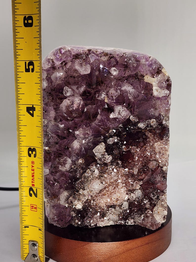 May include: A large amethyst crystal cluster, approximately 5 inches tall, mounted on a round wooden base. The crystal features a deep purple color with white and clear quartz inclusions. The base is a warm brown color.
