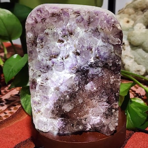May include: A rectangular amethyst crystal illuminated from within, mounted on a round, dark brown wooden base. The crystal features shades of purple, white, and brown. The base has a small black power cord.