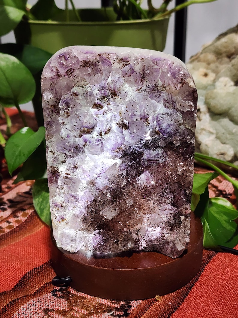 May include: A rectangular amethyst crystal illuminated from within, mounted on a round, dark brown wooden base. The crystal features shades of purple, white, and brown. The base has a small black power cord.