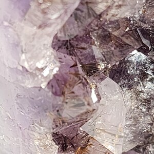 May include: Close-up of a cluster of amethyst crystals. The crystals display a range of colors, from deep purple to translucent white. The facets of the crystals catch the light, creating a sparkling effect. The overall texture is rough and natural.