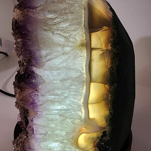 May include: A backlit amethyst geode lamp. The crystal formation features a gradient of purple, white, and amber hues. The lamp is mounted on a dark brown wooden base, creating a decorative and functional lighting piece.