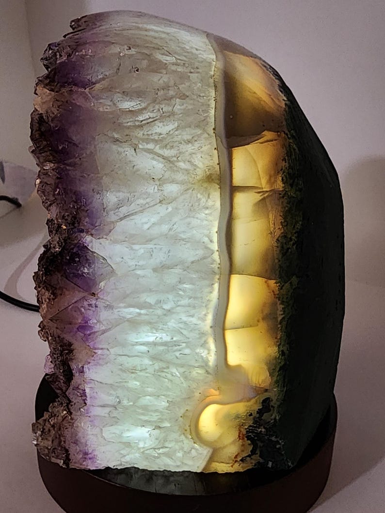 May include: A backlit amethyst geode lamp. The crystal formation features a gradient of purple, white, and amber hues. The lamp is mounted on a dark brown wooden base, creating a decorative and functional lighting piece.