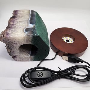 May include: A large, irregularly shaped amethyst geode with a green and white exterior. A wooden, round LED light base with a USB cable and on/off switch is next to the geode. The geode has a hollowed-out center.