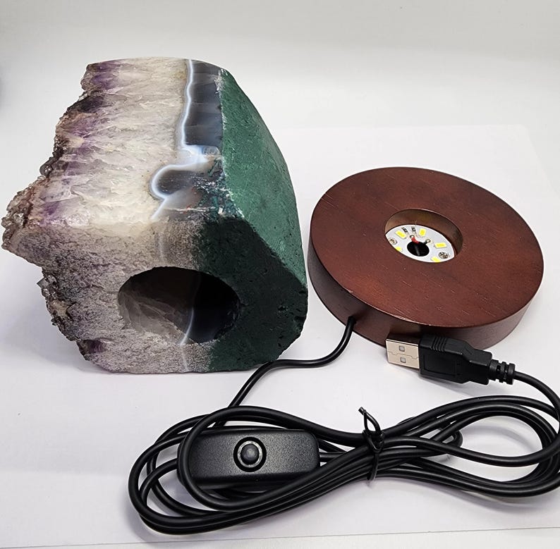 May include: A large, irregularly shaped amethyst geode with a green and white exterior. A wooden, round LED light base with a USB cable and on/off switch is next to the geode. The geode has a hollowed-out center.