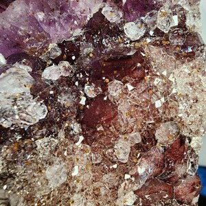May include: Close-up of a natural amethyst crystal cluster. The crystal features a deep purple hue with clear, sparkling quartz inclusions and a reddish-brown base. The surface is textured with various crystal formations.