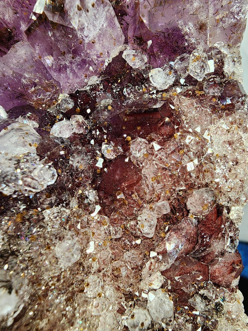 May include: Close-up of a natural amethyst crystal cluster. The crystal features a deep purple hue with clear, sparkling quartz inclusions and a reddish-brown base. The surface is textured with various crystal formations.
