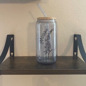 May include: A clear glass reusable water bottle with a bamboo lid and a black floral design. The bottle has the name "Maggie" written on it. A clear glass straw is inserted through the lid.