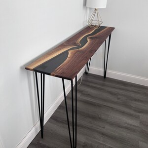 May include: A long, narrow console table with a black epoxy river flowing through a walnut wood top. The table is supported by four hairpin legs.