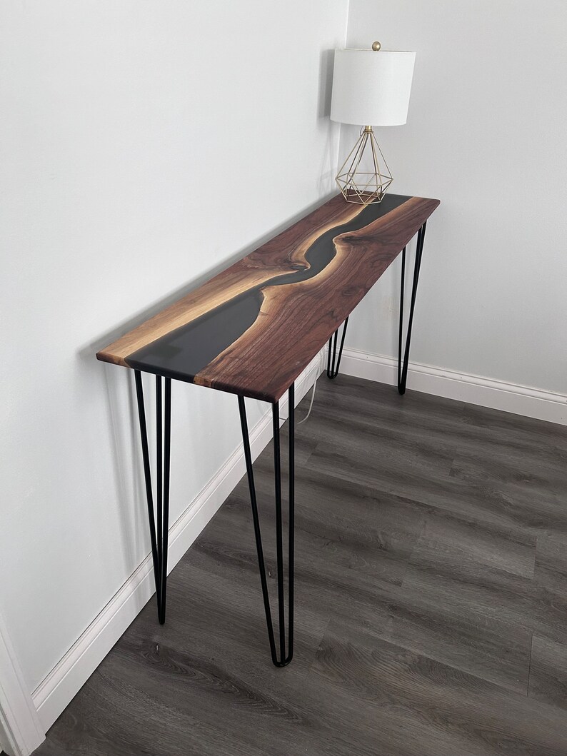 May include: A long, narrow console table with a black epoxy river flowing through a walnut wood top. The table is supported by four hairpin legs.