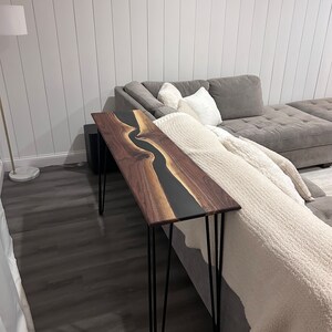 May include: A black and brown wooden console table with a black epoxy river design. The table has hairpin legs and is sitting in front of a gray couch.