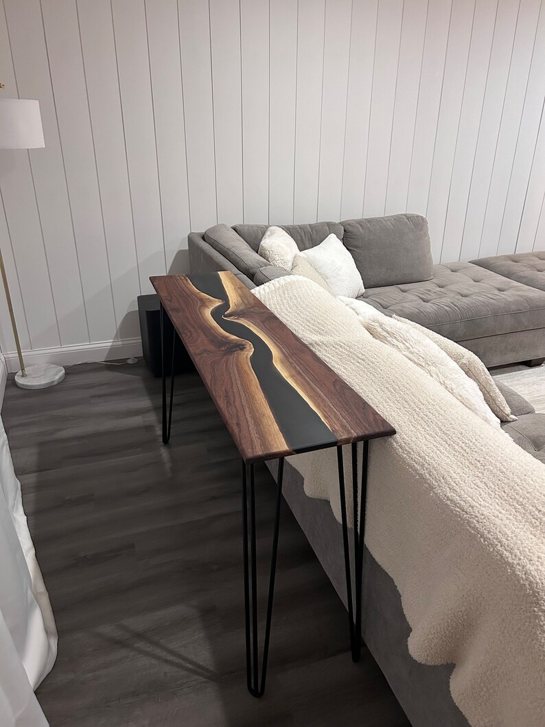 May include: A black and brown wooden console table with a black epoxy river design. The table has hairpin legs and is sitting in front of a gray couch.