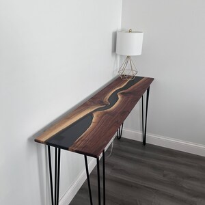 May include: A long, narrow console table with a black epoxy river flowing through a walnut wood top. The table is supported by four hairpin legs.