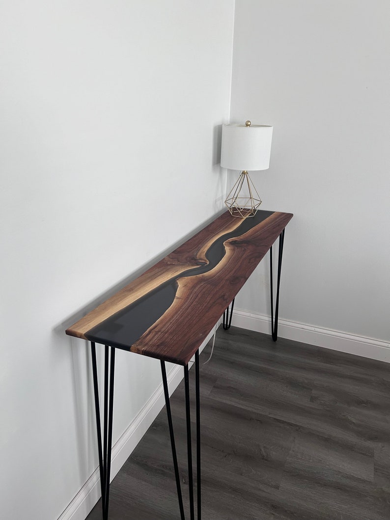 May include: A long, narrow console table with a black epoxy river flowing through a walnut wood top. The table is supported by four hairpin legs.