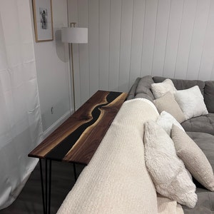 May include: A long, narrow walnut wood console table with a black epoxy river design. The table has hairpin legs and is placed next to a white couch with a white throw blanket and pillows.