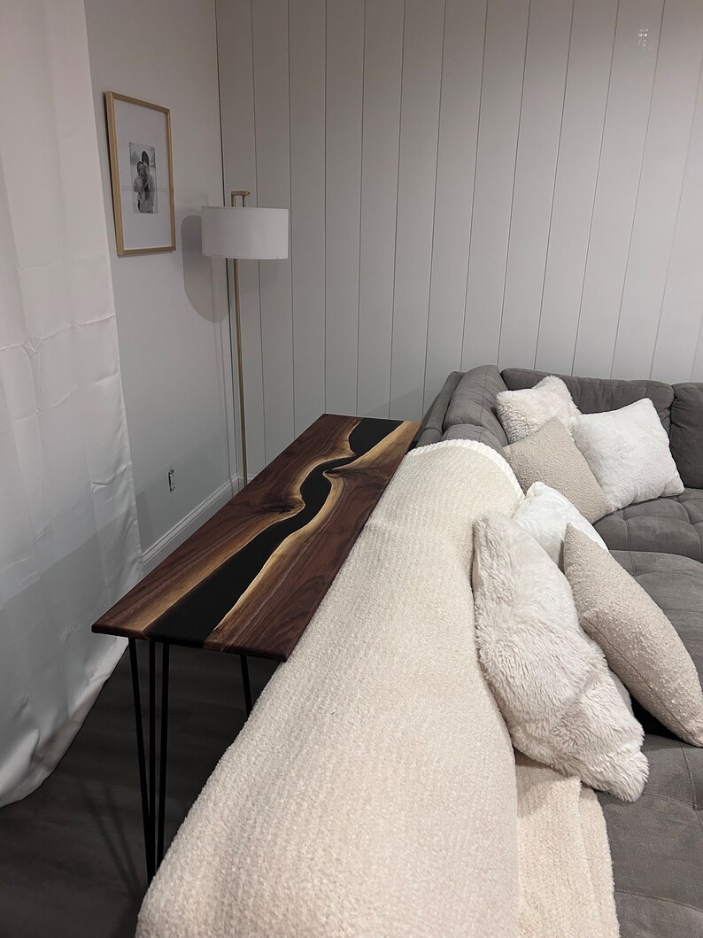 May include: A long, narrow walnut wood console table with a black epoxy river design. The table has hairpin legs and is placed next to a white couch with a white throw blanket and pillows.