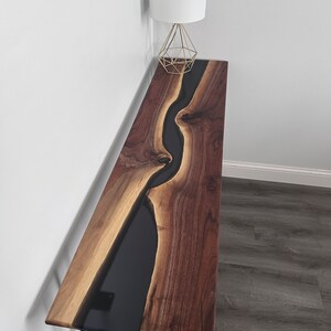 May include: A long, narrow console table with a black epoxy river flowing through the center of the walnut wood top. The table has black hairpin legs.