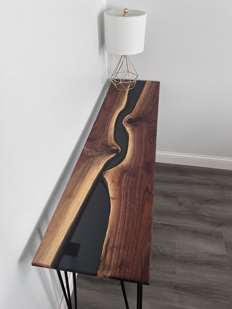 May include: A long, narrow console table with a black epoxy river flowing through the center of the walnut wood top. The table has black hairpin legs.