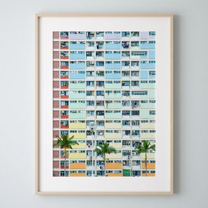 Op de afbeelding: Een ingelijste print van een kleurrijk appartementencomplex. Het gebouw heeft rijen balkons in pastelkleuren, waaronder blauw, groen, geel en oranje. Palmbomen zijn zichtbaar voor het gebouw.
