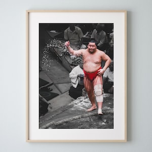 May include: A sumo wrestler in a red and white loincloth raises his arm in victory, covered in white powder. He is standing on a ring with a rope border, surrounded by spectators.