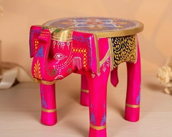 May include: A vibrant pink elephant-shaped wooden stool with intricate hand-painted details. The round top features a floral design with gold trim. The elephant's body is adorned with colorful patterns, making it a unique decorative piece.