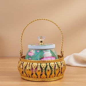 May include: A decorative gold-colored metal basket with a handle holds a teal and pink floral-patterned jar with a crystal knob. The jar is inside a gold filigree basket, creating an elegant display. The background is a warm neutral tone.