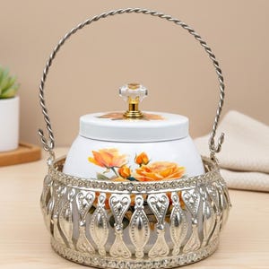 Könnte beinhalten: Ein weißer Keramikbehälter mit einem Blumendesign aus orangefarbenen Rosen, eingebettet in einen dekorativen silberfarbenen Metallkorb. Der Behälter hat einen Deckel mit einem Kristallknopf und einem silbernen Griff.