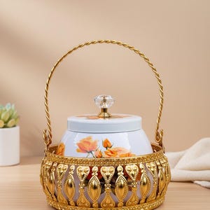 May include: A decorative white and gold serving dish with a floral design. The dish has a gold handle and a crystal knob on the lid. The base is gold with an ornate design. The dish is approximately 15 cm in diameter.