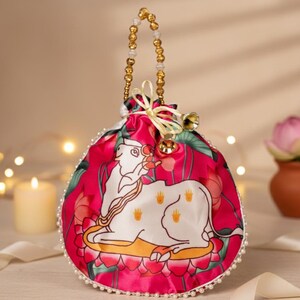 May include: A vibrant pink satin pouch with a white cow illustration and gold beaded handle. The pouch is adorned with a pearl trim and floral accents. A small gold bell hangs from the top, adding a touch of elegance.
