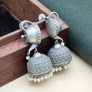 Op de afbeelding: Zilverkleurige oorbellen met een theepotontwerp en een hangende, klokvormige hanger met parel accenten.