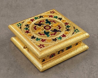 May include: A gold-colored, square jewelry box with an ornate floral design on the lid. The box features a detailed pattern around the edges and a central floral motif with red, green, and yellow accents. The box is open, revealing the interior.