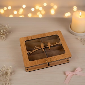May include: Wooden jewelry box with a clear top and a decorative bird branch design. The box is square, with a gold clasp, and is made of light brown wood. The interior is divided into compartments. A lit candle and a pink bow are in the background.