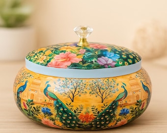 May include: A decorative, round jar with a lid. The jar is painted with a yellow background and features images of peacocks, trees, and buildings. The lid is adorned with colourful flowers and a crystal knob.
