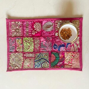 May include: A rectangular, hot pink, embroidered placemat with a patchwork design. The placemat features various embroidered patterns in gold, green, and blue. A small white bowl with a gold rim, filled with almonds, sits on the mat.