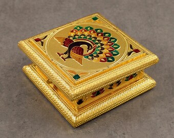 May include: A gold-colored, square jewelry box with a detailed peacock design on the lid. The peacock features a vibrant array of colors, including green, red, and yellow. The box has a textured, beaded border.