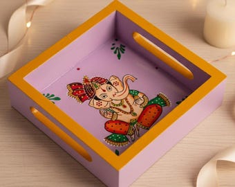 May include: Square wooden tray with a hand-painted image of the Hindu deity Ganesha. The tray has a lavender interior, yellow trim, and cut-out handles. The Ganesha illustration features vibrant colors and intricate details.