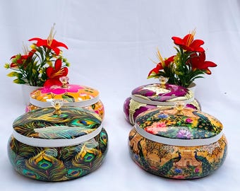 May include: Four decorative, round lidded jars with ornate designs. The jars feature vibrant colors and patterns, including peacock feathers and floral motifs. Each jar has a decorative knob on the lid. Red flowers in white pots are in the background.
