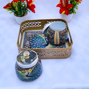 Peut inclure: Un plateau doré décoratif avec un motif de paon, contenant deux pots assortis avec des couvercles. Les pots et le plateau présentent un motif de paon bleu, vert et or. Deux petits pots blancs avec des fleurs rouges sont de chaque côté.